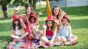 Two women and four girls enjoy a sunny picnic in Portugal, waving happily in the park.
